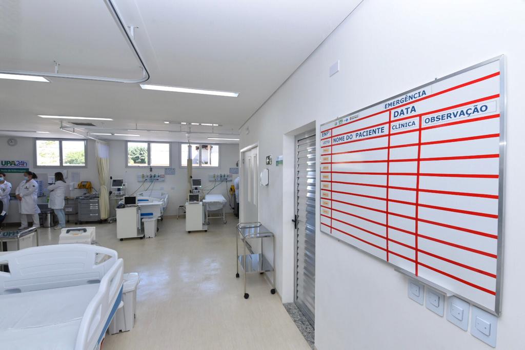Foto de clínica - A grande sala branca conta com camas de UTI e equipamentos médicos. Há três enfermeiras do lado esquerdo e na parede direita um quadro para informações dos pacientes